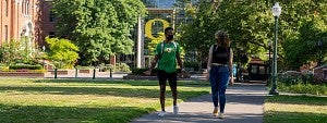 Two students wearing masks walking across campus near the O on Lillis Hall
