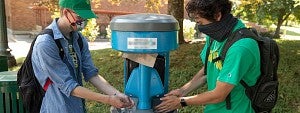 Two students wearing masks using an outdoor hand washing station on campus