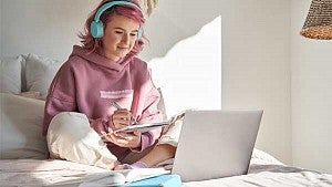 Girl sitting on her bed writing on a tablet while wearing headphones and working on a computer
